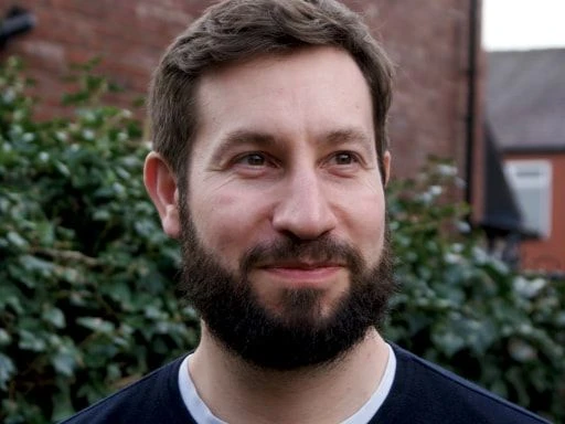 Bearded man smiling slightly while standing outside in front of a brick house and bushes.