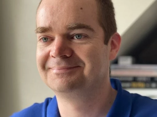Smiling man in a blue polo shirt indoors with books in the background.
