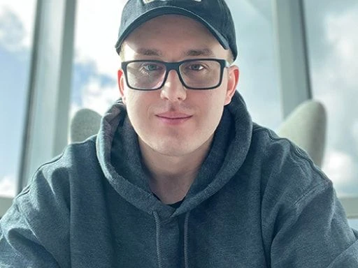 Young man wearing glasses, a dark hoodie, and a baseball cap, smiling slightly in front of large bright windows.