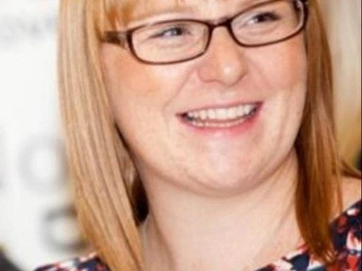 Woman with straight blonde hair, glasses, and a floral top smiling in a candid moment, appearing cheerful and engaged.
