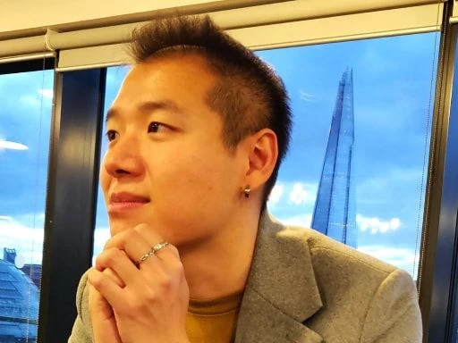 Man with short dark hair wearing earrings and rings, seated near a window with a modern city building in the background, looking contemplatively to the side.