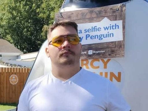 Man in a white t-shirt and yellow-tinted sunglasses posing in front of a sign that says “Take a selfie with Percy Penguin,” in a sunny outdoor setting.