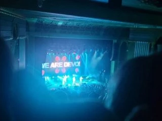 Brightly lit concert stage from an audience perspective, featuring the message 'We Are Devo!' on a large backdrop screen.