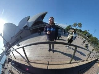 Fisheye lens photo of a man standing on the promenade outside the Sydney Opera House under a clear blue sky.